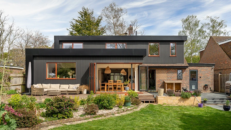 Rear view of a modern two-storey home with a black timber-clad extension, large sliding glass doors, landscaped garden, and outdoor seating area.