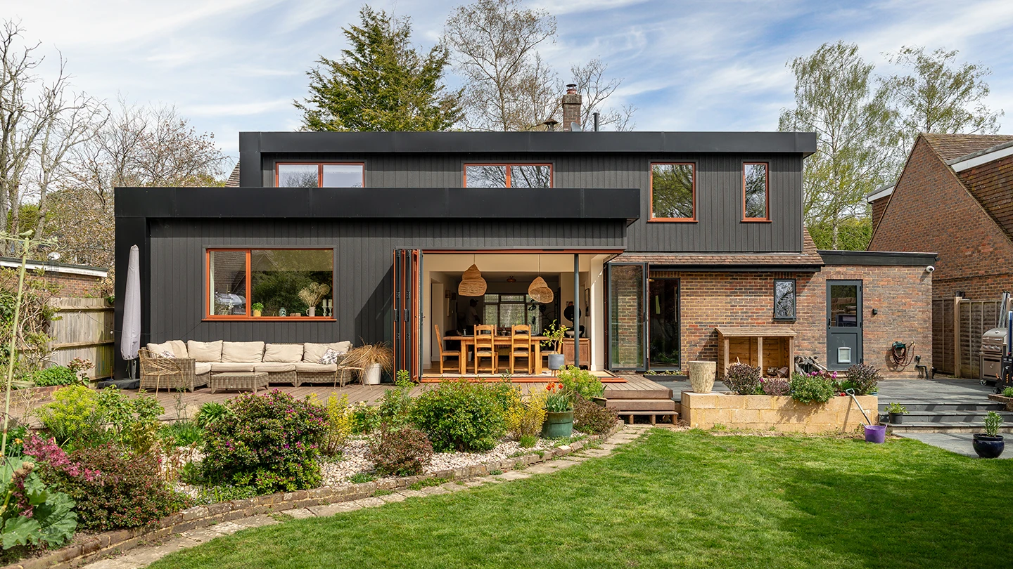 Rear view of a modern two-storey home with a black timber-clad extension, large sliding glass doors, landscaped garden, and outdoor seating area.